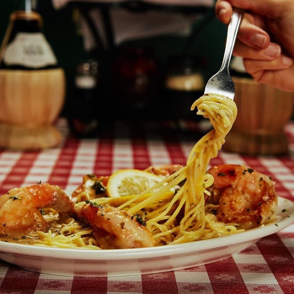 A fork lifts spaghetti with shrimp and lemon from a white plate on a red and white checkered tablecloth.