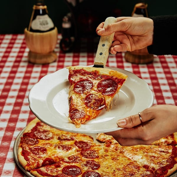 Hands serving a slice of Miceli's pepperoni pizza onto a white plate over a red checkered tablecloth.