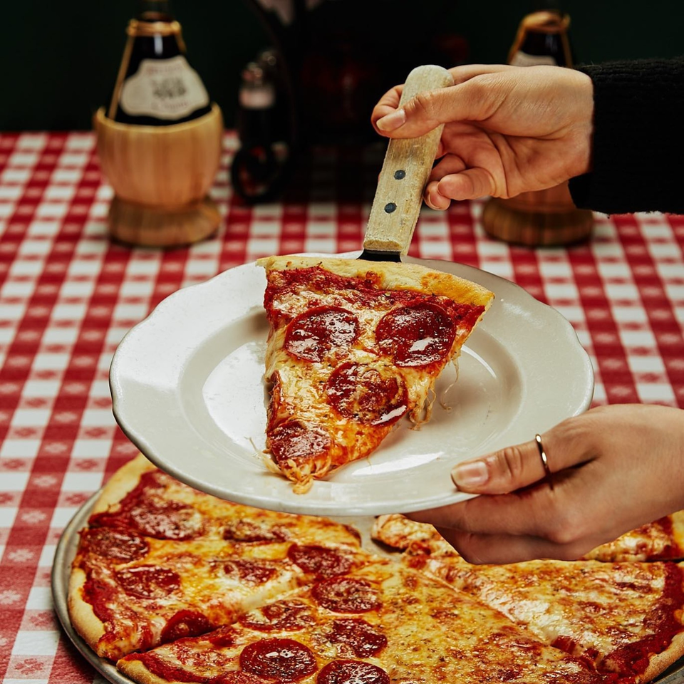 Hands serving a slice of Miceli's pepperoni pizza onto a white plate over a red checkered tablecloth.