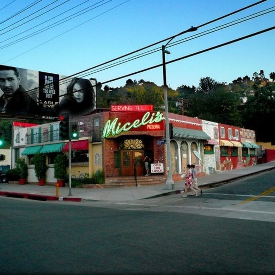 Exterior of Miceli's Pizzeria, a red brick building with green neon sign, on a sunny day.